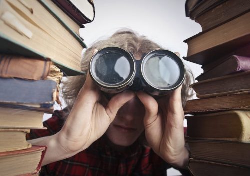 Young unrecognizable person sitting at table with stacked old books and looking through binoculars