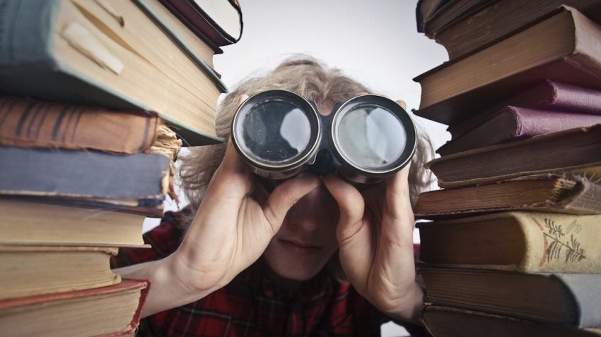 Young unrecognizable person sitting at table with stacked old books and looking through binoculars
